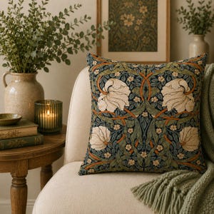 May include: A square throw pillow featuring a dark blue background with a repeating pattern of large cream-colored flowers, green foliage, and thin orange stems. The fabric has a woven texture and is placed on a light-colored upholstered chair.