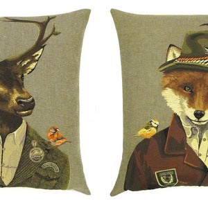 May include: Two decorative throw pillows with a beige background. One pillow features a deer wearing a suit and a hat with birds perched on its antlers. The other pillow features a fox wearing a suit and a hat with a bird perched on its shoulder.