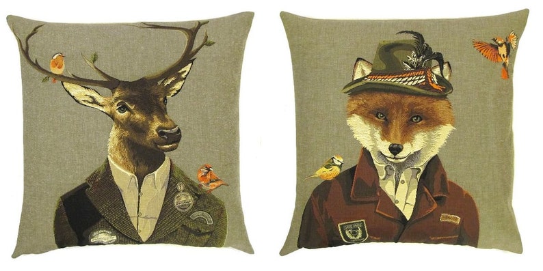 May include: Two decorative throw pillows with a beige background. One pillow features a deer wearing a suit and a hat with birds perched on its antlers. The other pillow features a fox wearing a suit and a hat with a bird perched on its shoulder.