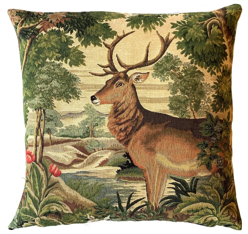 May include: A tapestry pillow featuring a brown deer with large antlers standing in a forest setting. The deer is surrounded by green trees and bushes, and there is a small stream in the background.