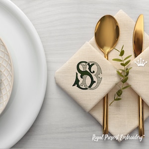 May include: A white linen napkin with a green embroidered monogram "SD" on it. The napkin is folded and placed on a table with gold silverware and a sprig of greenery.