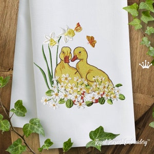 May include: Two yellow ducklings with orange beaks are nestled in a bed of white flowers with green leaves. The ducklings are facing each other and have their heads tilted. There are two orange butterflies in the background.