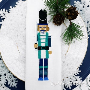 Puede incluir: Diseño de cascanueces bordado en una servilleta de lino blanco. El cascanueces lleva un uniforme azul y verde con un sombrero negro y sostiene una espada. El diseño está rodeado de un patrón de copo de nieve blanco.