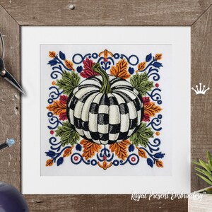 Puede incluir: Un dise&ntilde;o de bordado que presenta una calabaza central con un patr&oacute;n de cuadros en blanco y negro, rodeada de coloridas hojas de oto&ntilde;o en tonos naranja, rojo y verde, enmarcada por volutas decorativas azules sobre un fondo de tela blanca.