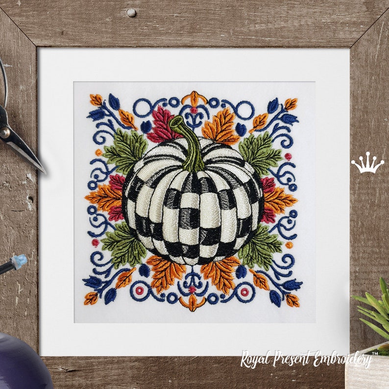 Puede incluir: Un dise&ntilde;o de bordado que presenta una calabaza central con un patr&oacute;n de cuadros en blanco y negro, rodeada de coloridas hojas de oto&ntilde;o en tonos naranja, rojo y verde, enmarcada por volutas decorativas azules sobre un fondo de tela blanca.