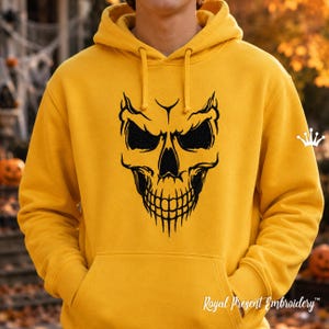 Könnte beinhalten: Ein senfgelber Kapuzenpullover mit einer schwarzen, gestickten Totenkopf-Grafik auf der Brust. Der Hoodie verfügt über eine Kängurutasche auf der Vorderseite, lange Ärmel und passende gelbe Kordelzüge am Halsausschnitt.