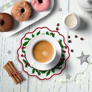 Op de afbeelding: Een witte koffiekop met schotel staat op een rood en groen geborduurde onderzetter met een hulstontwerp. De onderzetter staat op een witte houten tafel met een kan room, kaneelstokjes en donuts.