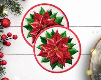Può includere: Due sottobicchieri rotondi ricamati di Natale con un fiore di stella di Natale rosso con foglie verdi su uno sfondo bianco con un bordo rosso.