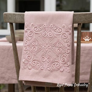 Op de afbeelding: Een oudroze linnen handdoek met een vierkant, toon-op-toon geborduurd patroon van symmetrische krullen en bloemmotieven. Het borduurwerk is gecentreerd op de stof, die over de rugleuning van een houten stoel is gedrapeerd.