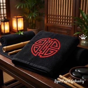 May include: Black spa towels with a red embroidered design are displayed on a wooden surface. Rolled towels, bamboo, and spa stones are also visible. The towels have a decorative design in the center.