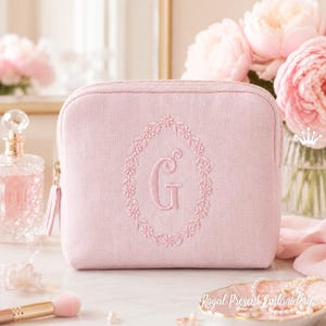 Peut inclure: Une trousse de maquillage rose clair avec un motif ovale brodé de fleurs et la lettre "G" au centre. Le sac a une fermeture éclair et un pompon. L'arrière-plan comprend un flacon de parfum, des fleurs et un pinceau de maquillage.