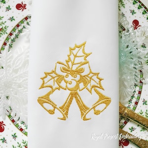 Puede incluir: Campanas de Navidad bordadas en oro con una rama de acebo y un lazo en una servilleta blanca. La servilleta está sobre un plato con un patrón navideño verde y blanco.