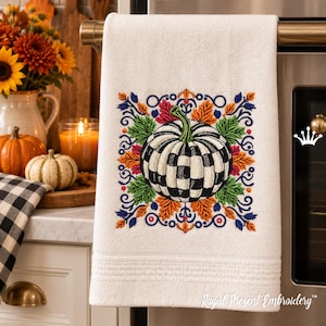 Puede incluir: Un pa&ntilde;o de cocina blanco con un dise&ntilde;o bordado de una calabaza a cuadros blancos y negros rodeada de coloridas hojas de oto&ntilde;o en tonos naranja, verde y azul, con el texto Royal Present Embroidery en la esquina inferior.