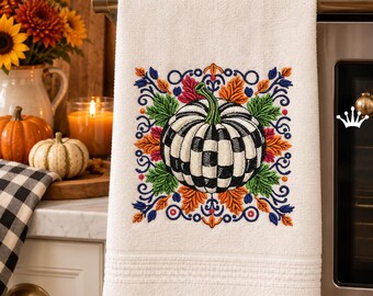 Pode incluir: Um pano de prato branco com um design bordado de uma abóbora xadrez preta e branca rodeada por folhas de outono coloridas em tons de laranja, verde e azul, com o texto Royal Present Embroidery no canto inferior.