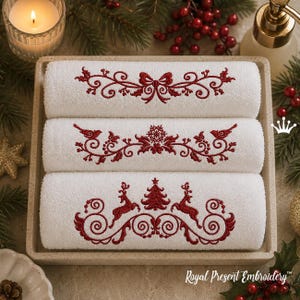 May include: Three white towels stacked in a square tray, each featuring detailed red embroidery. The top towel displays a bow, the middle towel shows birds and a snowflake, and the bottom towel depicts two reindeer flanking a Christmas tree.