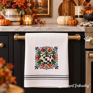 Puede incluir: Un pa&ntilde;o de cocina blanco con una calabaza bordada a cuadros blancos y negros, rodeada de coloridas hojas de oto&ntilde;o y adornos florales azules, con dos rayas grises horizontales en el borde inferior y el texto Royal Present Embroidery.