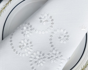 May include: White linen napkin with a delicate embroidered design of a stylized heart shape. The napkin is folded and placed on a white plate with a blue rim. The text "Royal Present Embroidery" is visible in the bottom right corner.
