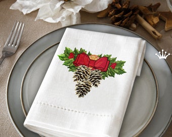 Könnte beinhalten: Eine weiße Leinen-Serviette mit einem gestickten Design aus Tannenzapfen, Stechpalmenblättern und einer roten Schleife. Das Design ist mittig auf der Serviette platziert und von einem gepunkteten Rand umgeben.