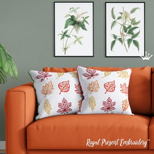 Könnte beinhalten: Zwei weiße Kissen mit gestickten Herbstblättern in Rot-, Orange- und Gelbtönen. Die Kissen liegen auf einem orangen Sofa vor einer grünen Wand mit zwei gerahmten botanischen Drucken.