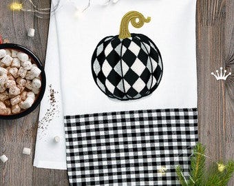 May include: A white kitchen towel with a black and white checked border. The towel features an embroidered black and white pumpkin with a gold stem.