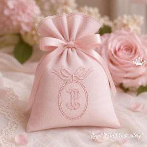 Puede incluir: Una bolsa rosa claro con cordón y cierre de cinta de raso. La bolsa presenta un diseño ovalado bordado con la letra "P" y un lazo. Las palabras "Royal Present Embroidery" están impresas en la parte inferior.