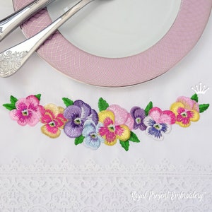 Op de afbeelding: Borduurwerk van een rij kleurrijke viooltjes met groene bladeren op een witte stoffen achtergrond. Het ontwerp is omlijst door een witte kantboord. De tekst "Royal Present Embroidery" is zichtbaar onderaan de afbeelding.