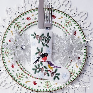 May include: White fabric Christmas stocking with embroidered pine branches, pine cones, and two birds. The stocking is on a white plate with a green and red holiday design.