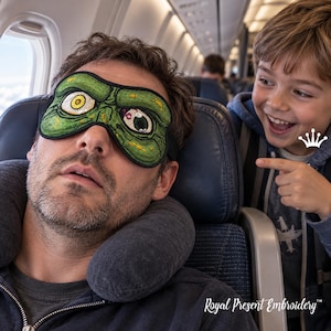 May include: A green sleep mask featuring an embroidered monster face with two distinct eyes, one yellow and one white with a black pupil, and textured skin details. The mask is worn over the eyes, covering the upper face area completely.