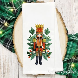 May include: Embroidered kitchen towel featuring a colorful nutcracker soldier with a red and gold uniform, a crown, and a mustache. The nutcracker is surrounded by green holly leaves and red berries.
