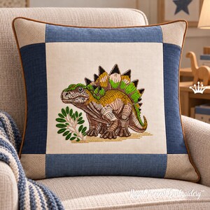 Op de afbeelding: Een vierkant sierkussen met een centraal crèmekleurig paneel met een geborduurde Stegosaurus-dinosaurus in bruine en groene tinten, omringd door een blauwe en beige rand met bruine biezen langs de buitenranden.