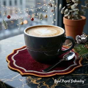 Puede incluir: Una taza de café gris oscuro con un diseño en forma de corazón en la espuma, sobre un posavasos burdeos con ribete dorado. Una cuchara plateada descansa junto a la taza. La escena está ambientada cerca de una ventana con luces y decoraciones festivas.