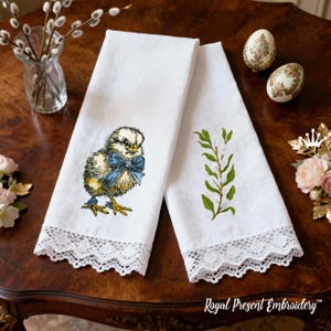 May include: Two white linen towels with embroidered Easter designs. One towel features a chick with a blue bow, the other has a sprig of pussy willow. Both towels have a white lace trim. The towels are on a dark wooden table.