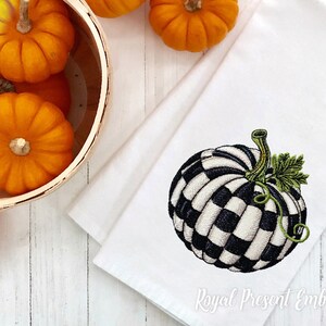 May include: A white kitchen towel featuring an embroidered black and white checkered pumpkin with a green stem and leaf. The towel is positioned next to a wooden basket filled with small orange pumpkins on a white wooden surface.