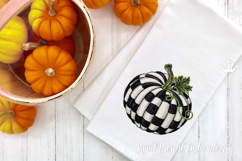 May include: A white kitchen towel featuring an embroidered black and white checkered pumpkin with a green stem and leaf. The towel is positioned next to a wooden basket filled with small orange pumpkins on a white wooden surface.