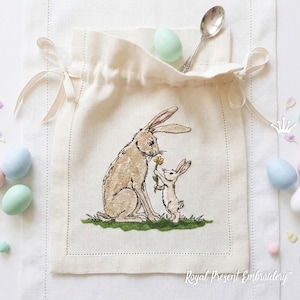 May include: A cream-colored drawstring bag with an embroidered design of two rabbits, one offering a flower. The bag has a pocket with a silver spoon and a light green egg. Pastel eggs and confetti surround the bag.