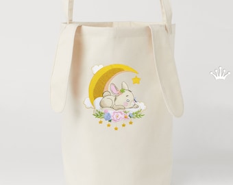 May include: A cream-colored canvas storage bin with long bunny ear handles. The bin features an embroidered design of a bunny sleeping on a cloud with a crescent moon and stars.