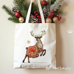 May include: A white tote bag featuring an embroidered reindeer with gold antlers and a red and gold patterned blanket. A gold snowflake is next to the reindeer. The bag is decorated with Christmas ornaments and greenery.