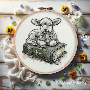 May include: Embroidered artwork in a wooden hoop depicts a lamb resting on a book with a cross. The lamb is white and grey, and the book is dark green with gold-coloured pages. The embroidery is surrounded by flowers and the text "Royal Present Embroidery".