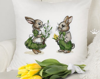 Peut inclure: Un coussin brodé représentant deux lapins tenant des bouquets de fleurs. Le lapin à gauche porte une robe verte et le lapin à droite porte un costume vert. Le coussin est blanc avec une texture subtile.