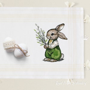 May include: Embroidered design of a brown bunny rabbit holding a sprig of willow branches. The bunny is wearing green overalls and a white shirt. The design is on a white background with a subtle plaid pattern.