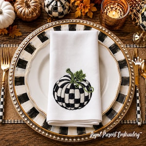 May include: A white cloth napkin featuring an embroidered black and white checkered pumpkin with a green stem and leaf. The napkin is folded and placed on a white dinner plate, which sits atop a black and white striped charger plate.