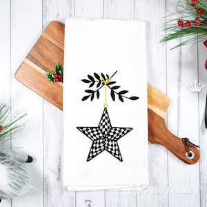 May include: A white kitchen towel with a black and white checkered star ornament hanging from a black twig with green leaves. The ornament is hanging from a gold string. The text "Royal Present Embroidery" is at the bottom of the towel.