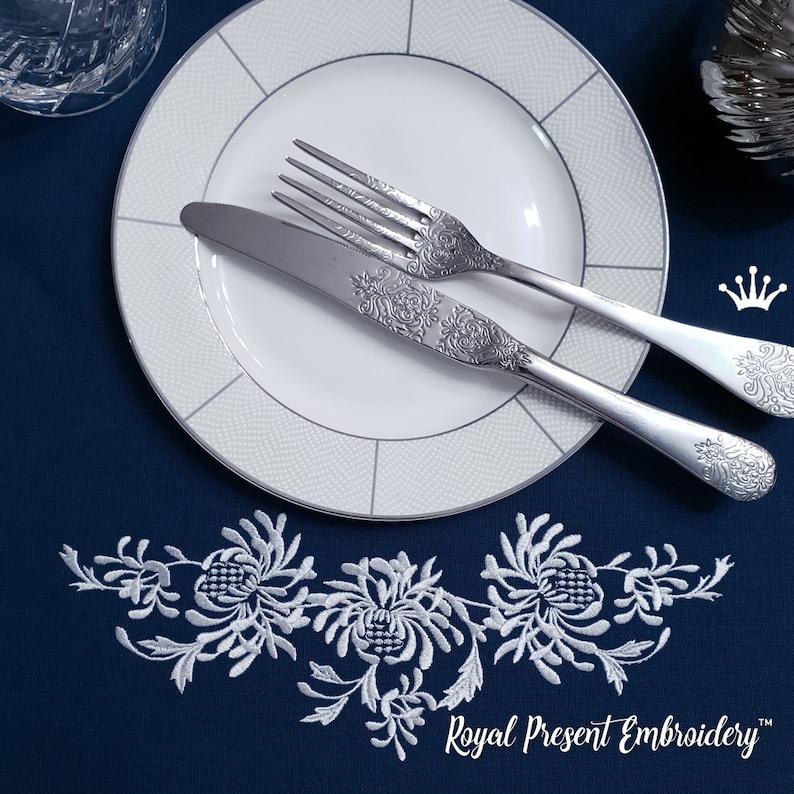 May include: Elegant table setting featuring a white plate with a silver rim, a fork, and a knife with ornate floral detailing. A white embroidered floral design adorns the navy blue tablecloth. The text "Royal Present Embroidery" is visible.