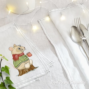May include: White linen napkins with a black stitched border. One napkin features an embroidered illustration of a mouse wearing a red scarf and green sweater, holding a mug. Silverware and fairy lights are also visible.