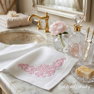 May include: White linen hand towel with pink floral embroidery. A clear glass bottle with a crystal stopper and a glass of toothbrushes sit on a marble countertop. A bar of soap and a pink flower are also present. The text "Royal Present Embroidery" is visible.