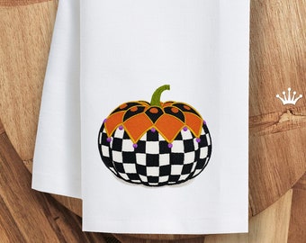 May include: A white kitchen towel with an embroidered black and white checked pumpkin with orange and gold accents. The pumpkin has purple beads around the top.