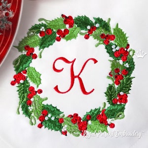 Puede incluir: Corona de Navidad bordada con bayas rojas y hojas verdes de acebo, con la letra K en el centro. La corona está rodeada de un fondo blanco con el texto "Royal Present Embroidery" en la parte inferior.
