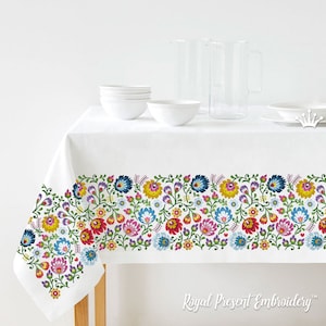 May include: White tablecloth with a colorful floral embroidery border. The embroidery features a repeating pattern of flowers and leaves in shades of red, yellow, blue, green, and pink. The tablecloth is set on a table with two clear glass pitchers and several white bowls.