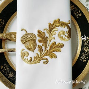 May include: White napkin with gold embroidery featuring an acorn and ornate leaves. The napkin is placed on a decorative plate with gold trim and a gold fork and knife. The text "Royal Present Embroidery" is at the bottom.