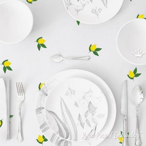 May include: A white tablecloth with a gray and white floral design on the plates. The tablecloth is decorated with embroidered lemons and green leaves. There are also two white bowls and a silver fork and knife on the table.